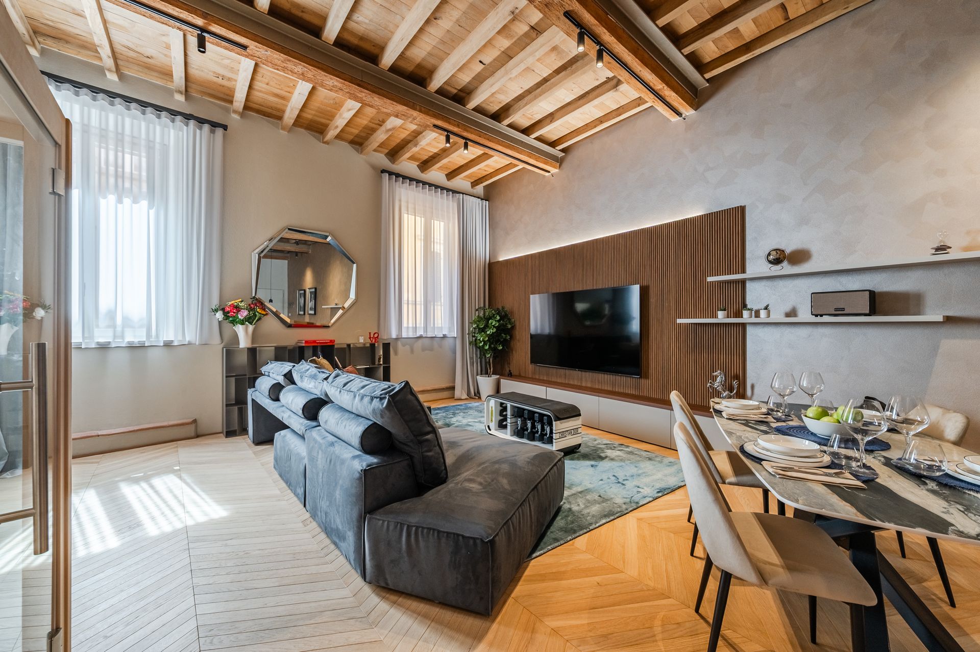 Salotto completo con divano blu-grigio, parete TV a doghe di legno, tavolo apparecchiato e travi a vista
